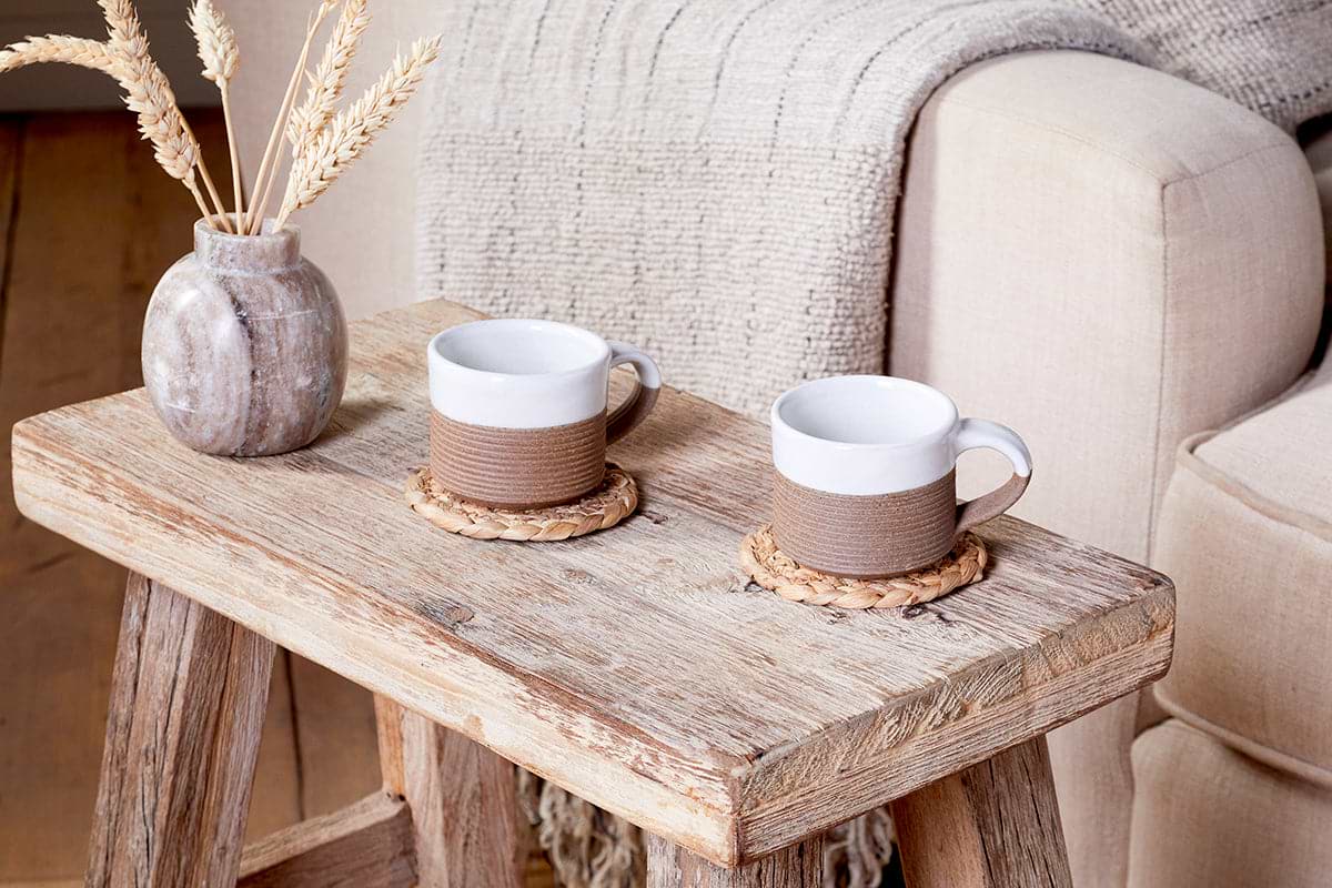 A rustic wooden coffee table displays nkuku's Mali Ribbed Espresso Mugs - White (Set of 2) on woven coasters, beside a ceramic vase with dried wheat. A beige sofa and textured blanket create a warm, inviting setting.