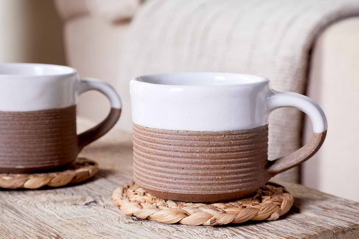 The nkuku Mali Ribbed Espresso Mugs in white (set of 2) rest on woven coasters atop a rustic wooden table, while a soft beige blanket drapes over a sofa in the background, creating a cozy atmosphere.