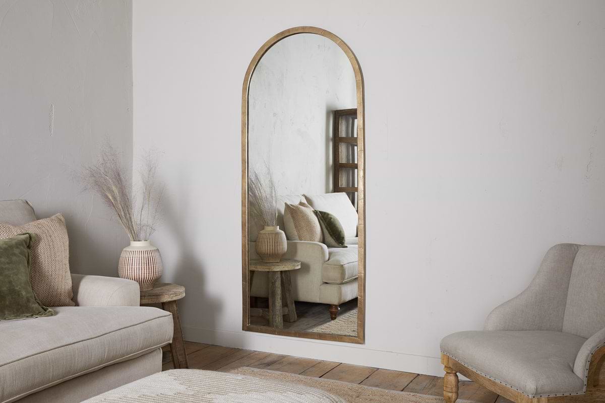 A neutral-toned living room features the nkuku Murwara Full Length Mirror in Antique Brass, reflecting a white sofa with green pillows, a side table, and a vase with dried branches. Wooden floors and light furniture create a calm, cozy atmosphere.