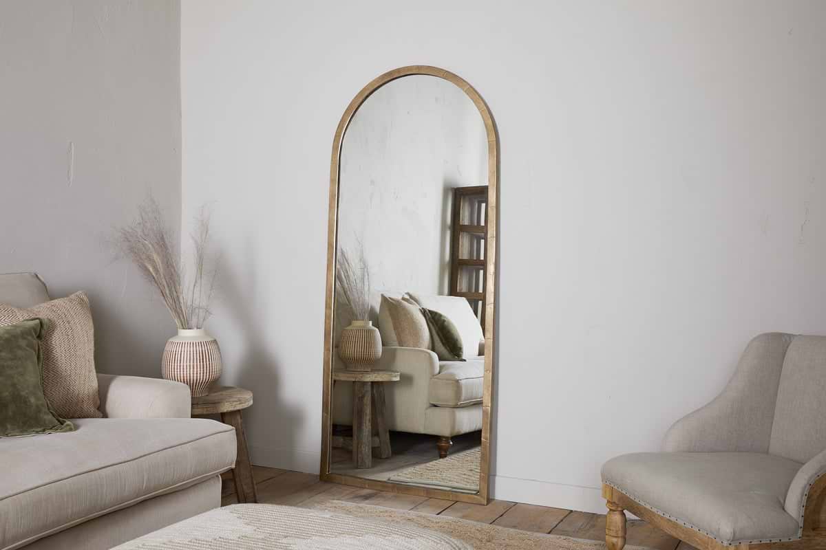 The nkuku Murwara Full Length Mirror in Antique Brass leans against a white wall, reflecting a beige sofa, green pillow, and décor. A wicker vase with dried grass sits on a wooden stool beside light wood floors and chairs.