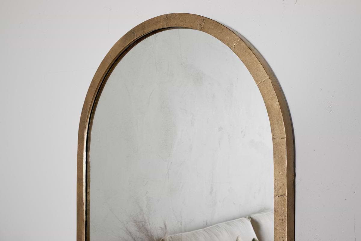 The Murwara Full Length Mirror by nkuku, featuring an arched antique brass frame, hangs on a white wall. Its reflection shows a light sofa and subtle decor, adding minimalist elegance to the space.