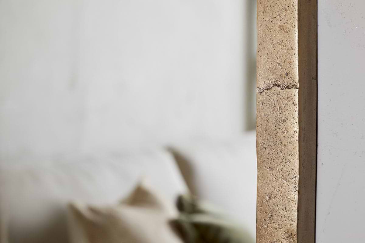 Close-up of a textured beige concrete wall with a visible crack. In the blurred background, the nkuku Murwara Full Length Mirror in Antique Brass stands beside a white sofa and neutral pillows, creating a soft, minimalist interior.