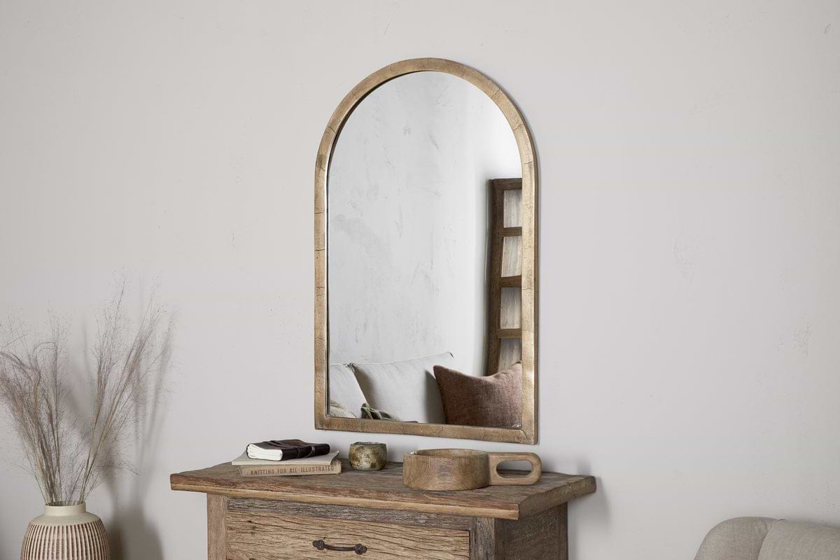 The nkuku Murwara Wall Mirror in antique brass with an arched wooden frame hangs above a rustic table with books, a candle, and a tray. A vase of dried grasses sits to the left, while the cozy sofa is reflected in the mirror.