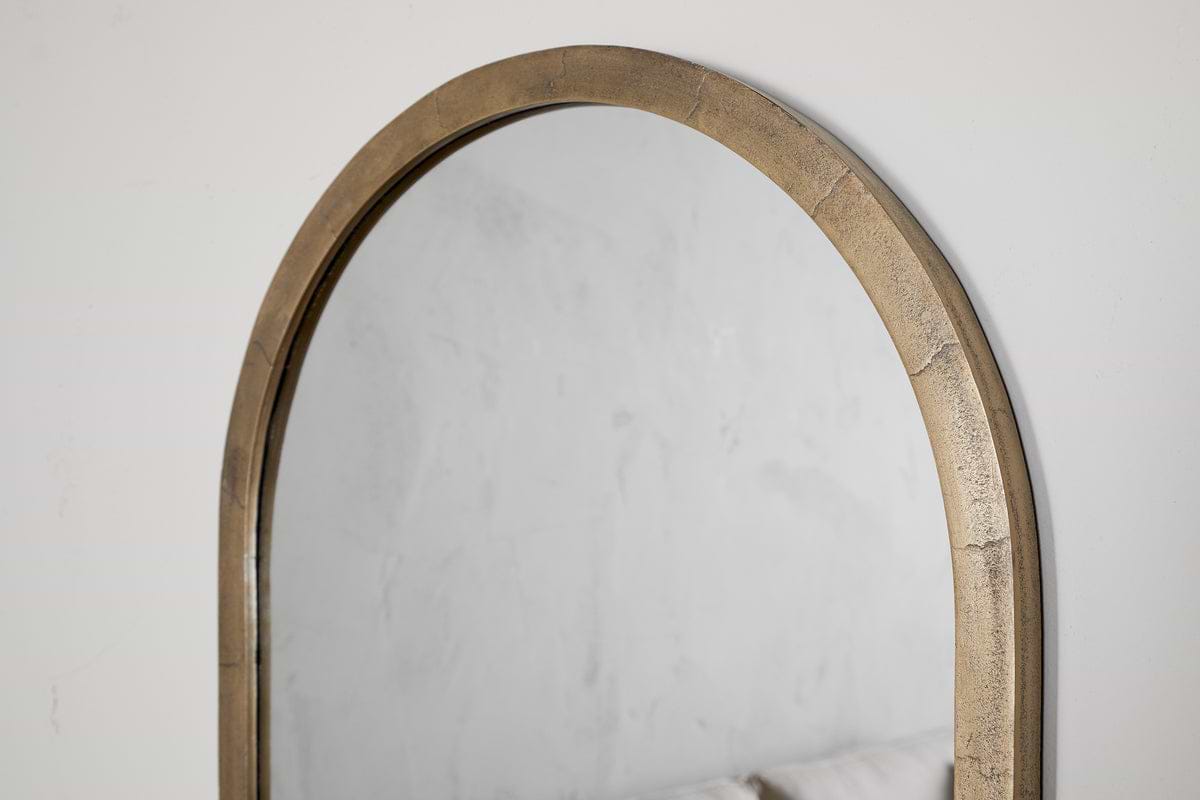 A close-up of the nkuku Murwara Wall Mirror in Antique Brass, reflecting a light-textured wall and the upper edge of a pale sofa.