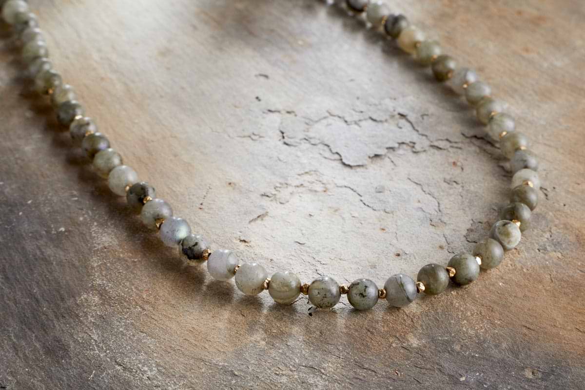 The Marai Necklace - Labradorite by nkuku showcases round, translucent grey-green labradorite stones and small gold spacer beads, elegantly arranged on a textured surface—an exquisite piece that embodies natural beauty.