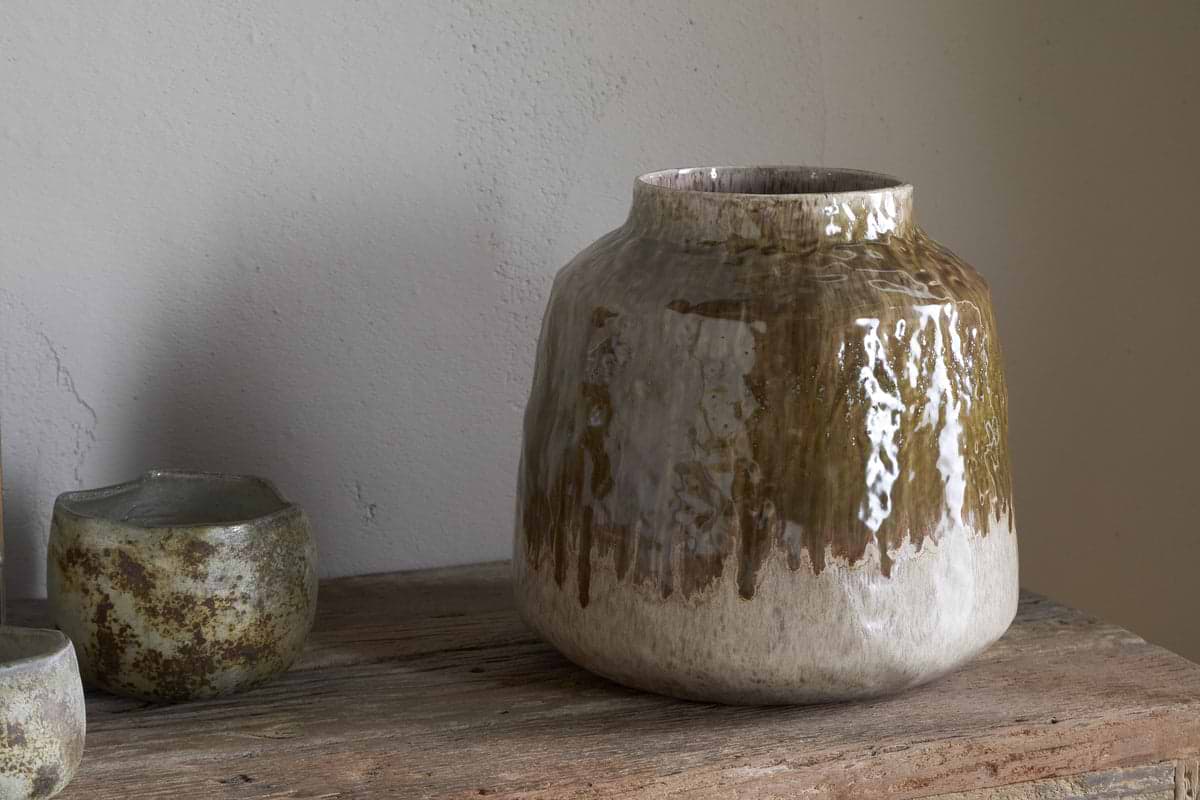 The nkuku Melina Reactive Glaze Vase in Olive & Natural sits on a rustic wooden surface beside two small textured ceramic cups, set against a plain light-colored wall.