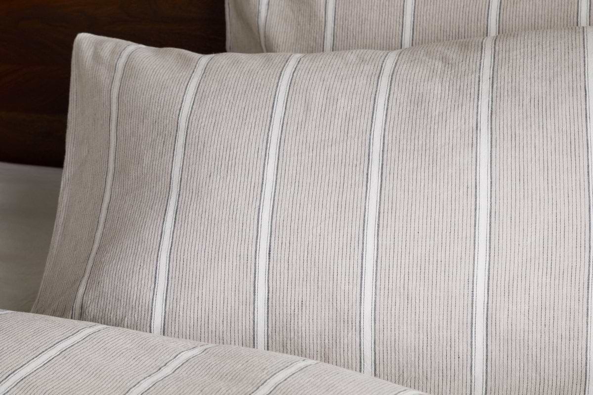 Close-up of nkuku’s Mora Hemp & Cotton Housewife Pillowcases in Stone Grey featuring thin vertical stripes and a soft, textured fabric. A matching pillow and part of a blanket appear in the background.
