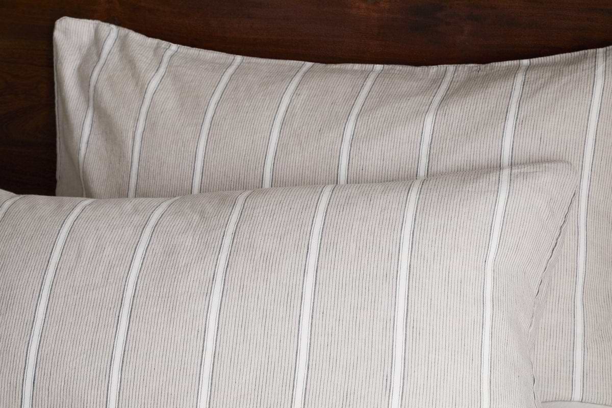 Two pillows with Mora Hemp & Cotton Housewife Pillowcases in Stone Grey by nkuku rest against a dark wooden headboard, featuring soft, comfortable fabric with subtle thin stripes.