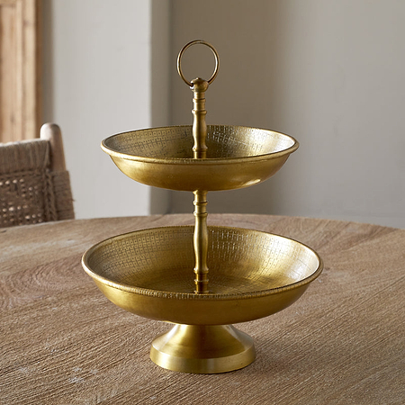 The Mahika Stand - Antique Brass by nkuku, a two-tiered gold metal serving tray ideal as a centerpiece or cake stand, is displayed on a round wooden table with woven chairs in the background.