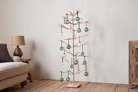 The nkuku Mala Base Jute Tree - Natural, with minimalist jute branches and gold accents, is adorned with green and clear glass ornaments, standing beside a beige sofa, rustic wooden cabinet, and table lamp against an off-white wall.