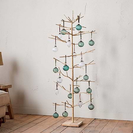 The nkuku Mala Base Jute Tree - Natural, with minimalist jute branches and gold accents, is adorned with green and clear glass ornaments, standing beside a beige sofa, rustic wooden cabinet, and table lamp against an off-white wall.