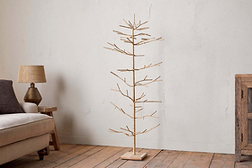 The nkuku Mala Base Jute Tree - Natural, with woven jute branches and a wooden base, stands on a wooden floor next to a beige sofa and small lamp table, set against a plain white wall in this cozy, rustic living room.