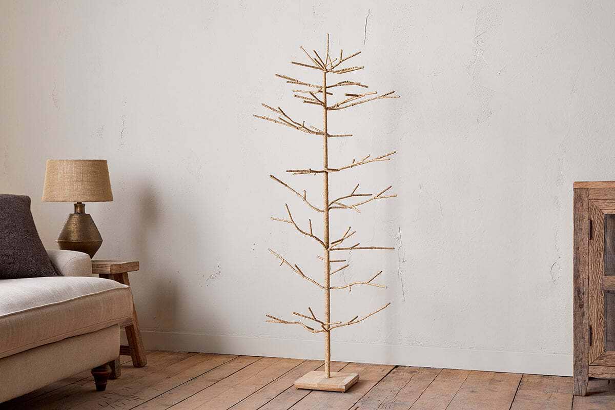 The nkuku Mala Base Jute Tree - Natural, with woven jute branches and a wooden base, stands on a wooden floor next to a beige sofa and small lamp table, set against a plain white wall in this cozy, rustic living room.