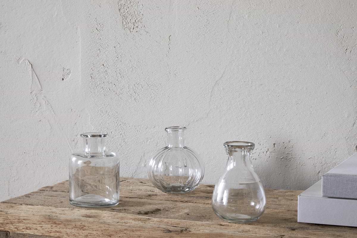 The nkuku Mayumi Recycled Glass Bud Vases – Clear (Set of 3) features three empty, uniquely shaped vases arranged on a rustic wooden surface against a textured off-white wall, with closed white books partially visible nearby.