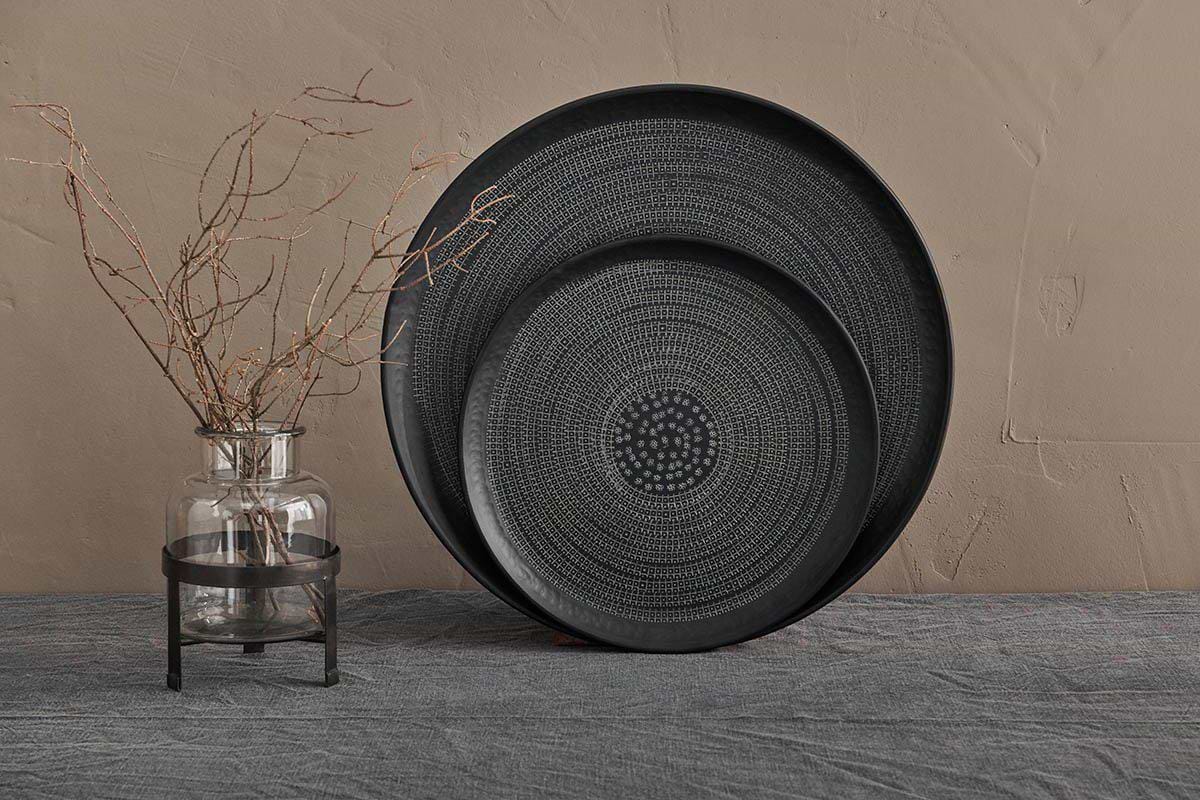 A clear glass vase with thin branches sits on a gray textured cloth beside two overlapping nkuku Mahika Trays in black, featuring hand-etched, intricate dotted patterns, all set against a subtly textured beige plaster wall.
