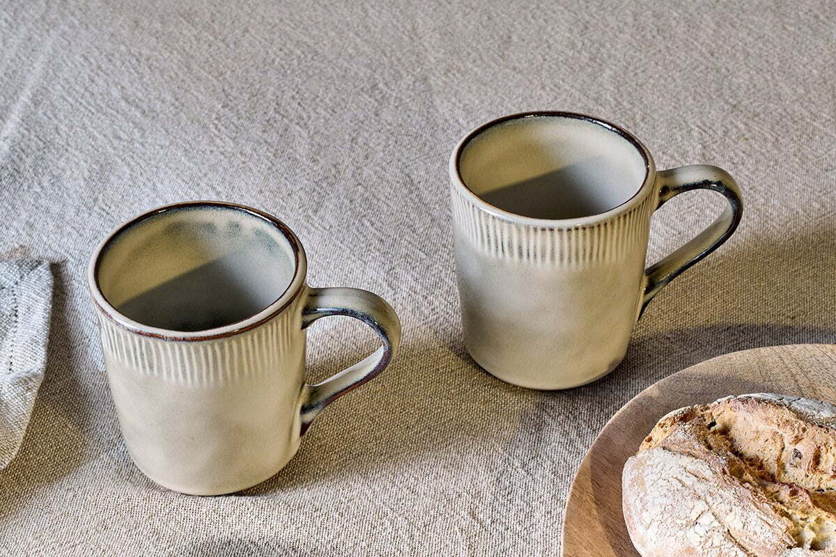 Two nkuku Malia Mug - Cream (Set of 2) with ribbed texture and dark brown rims sit on a linen tablecloth, with a rustic loaf of bread on a wooden board visible in the lower right corner.