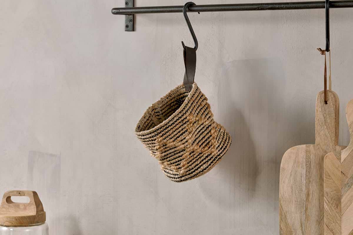 The nkuku Mannu Cotton & Hemp Wall Hung Basket in Natural & Black hangs from a black metal hook on a wall-mounted rod, with light wooden cutting boards leaning nearby and a glass jar with a wooden lid sitting to the left.