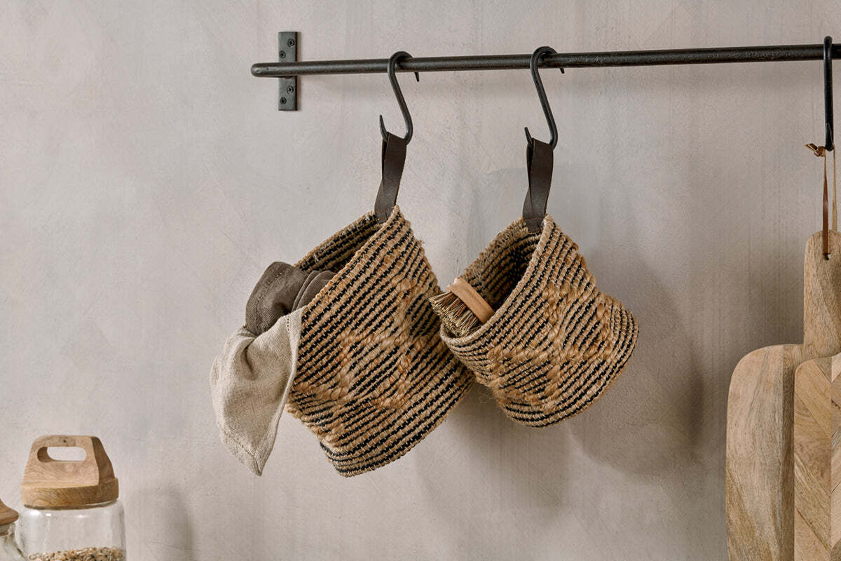 Two nkuku Mannu Cotton & Hemp Wall Hung Baskets in Natural & Black, suspended by leather straps from black hooks, store a beige cloth and wooden utensils beside glass jars and cutting boards.