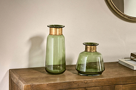 Two nkuku Miza Glass Vases in green, one tall and cylindrical, the other short and wide with copper rims, sit on a wooden surface. A round mirror and stacked books appear in the background against a light wall.