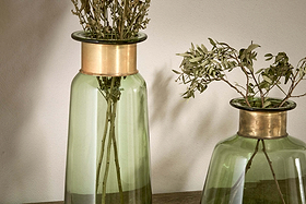 Two Miza Glass Vase - Green by nkuku, each with a wide gold rim, display dried leafy branches. Their simple, modern design and green tinted glass stand out on a wooden surface against a plain light wall in soft lighting.