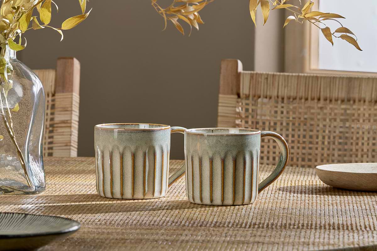 The nkuku Moda Mug - Natural (Set of 2), featuring a ribbed texture and earthy glaze, sits on a woven table. Wooden chairs with cane backs, dried leaves, and warm sunlight create a cozy background.