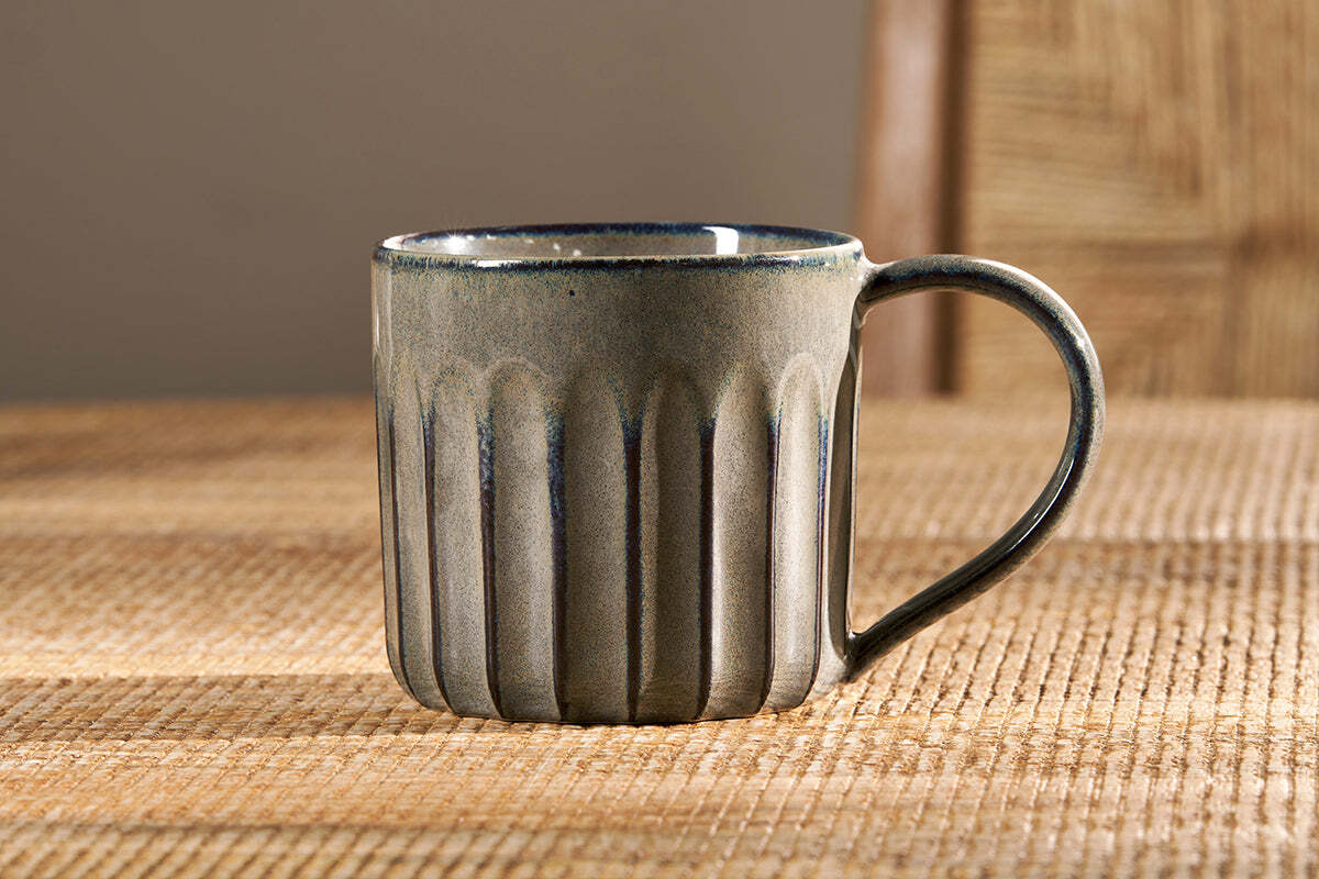 The nkuku Moda Mug - Pewter (Set of 2) features vertical ribbing and a blue-grey pewter glaze, showcased beautifully on a woven placemat to highlight its elegant grooved design.