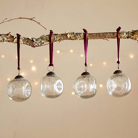 A bare branch with moss and lichen displays four nkuku Nishan Pinched Baubles - Clear, each hung by burgundy velvet ties. Soft fairy lights glow in the background, creating an elegant and festive beige-wall display.
