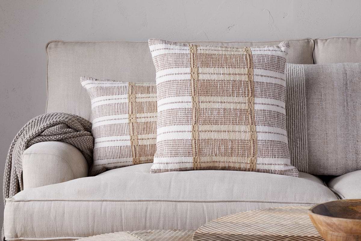 Two Nayika Cotton Cushion Covers by nkuku in rust add warmth to a light tan sofa, with a knitted throw over the armrest. The neutral setting and cosy decor create an inviting atmosphere.