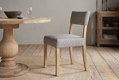 The Nadeeka Linen Upholstered Dining Chair in Soft Grey by nkuku, with a cushioned seat and upholstered backrest, sits on a woven round rug next to a wooden dining table. A wooden sideboard and glasses of white wine are visible in the background.