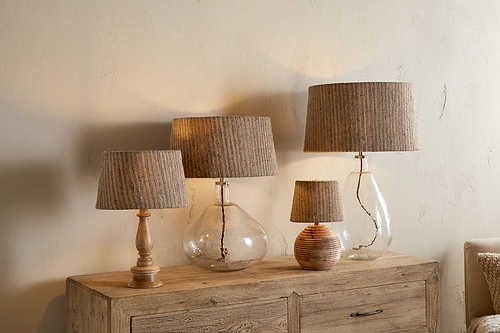 Four nkuku Nadee Banana Leaf Lampshade table lamps, featuring natural textured shades and bases of clear glass and wood, are arranged on a wooden sideboard against a beige wall, creating a cozy, rustic ambiance.