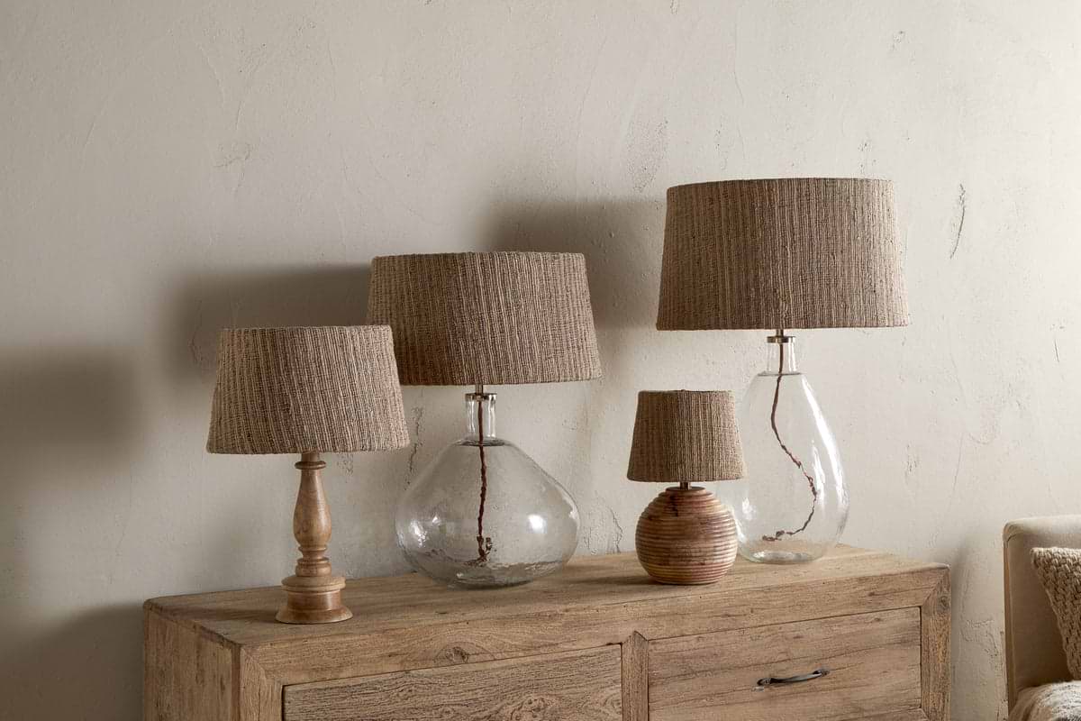 Four Nadee Banana Leaf Lampshades by nkuku, featuring natural textured shades atop glass or wooden bases, are arranged on a rustic wooden sideboard against a neutral wall, creating a cozy and organic look.