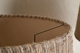 Close-up of the nkuku Nadee Banana Leaf Lampshade in natural tones, featuring woven texture and a visible metal support arm inside. The background displays a subtly textured beige wall.