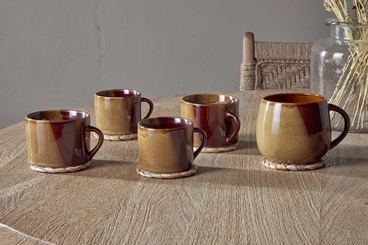 Two nkuku Naav Mugs - Mustard (Large) sit on woven coasters atop a wooden table, with a rustic chair and a glass vase filled with dried plants in the background.