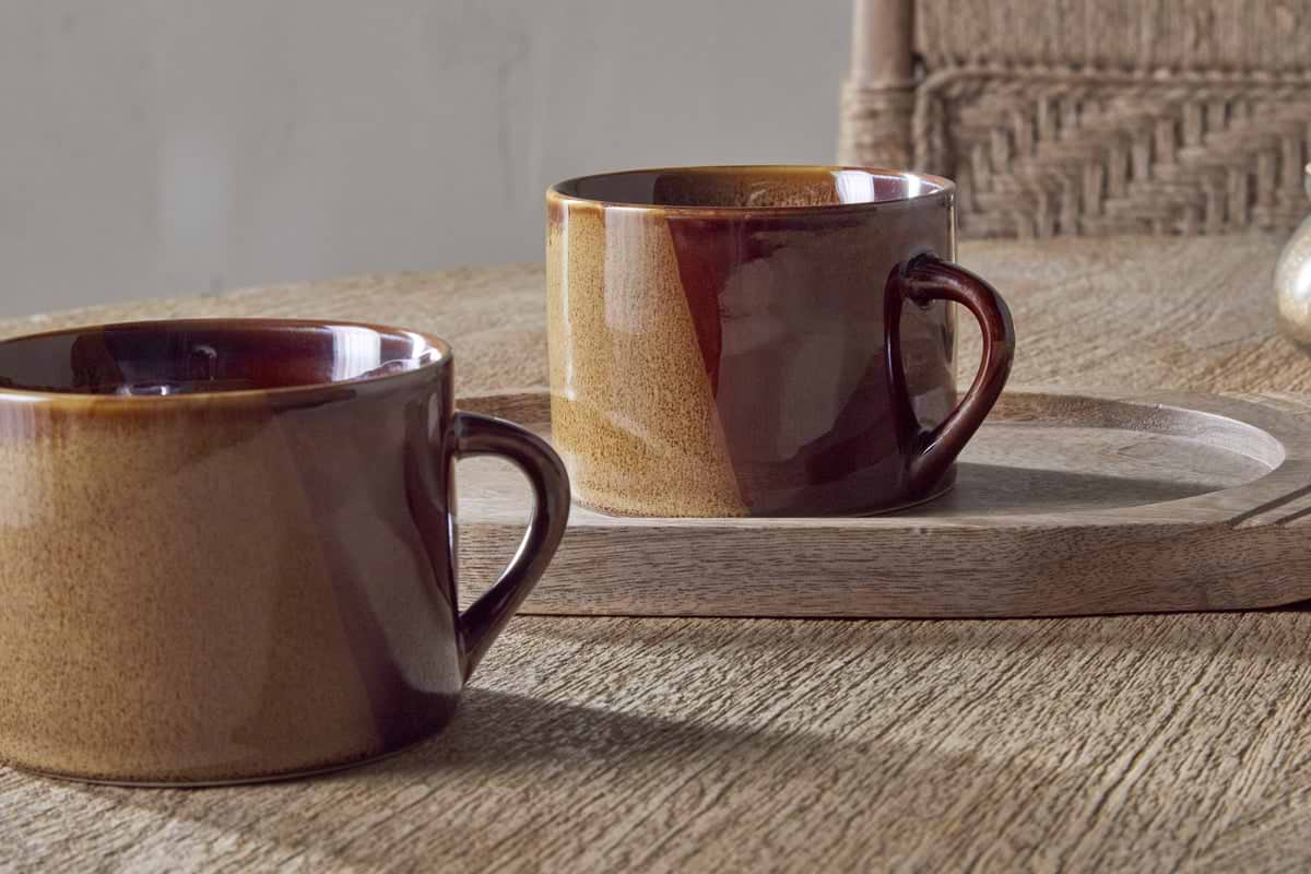 Two Naav Mugs in mustard, large size from nkuku, each with a unique glaze effect, sit on a wooden surface; one is on a round tray and a woven chair is in the background.