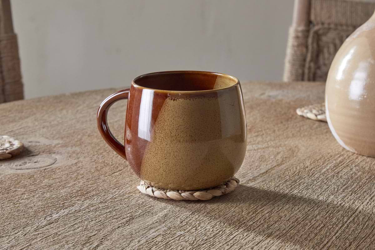 The Naav Mug - Mustard (Extra Large) by nkuku sits on a round woven coaster on a wooden table, with part of a vase and two woven placemats in the background.