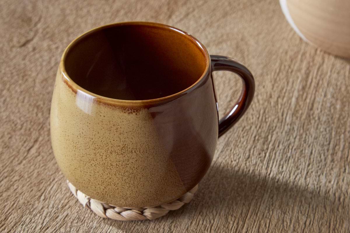 The Naav Mug - Mustard (Extra Large) by nkuku is a round ceramic mug with a brown and beige reactive glaze, paired with a woven coaster on a textured wooden surface.