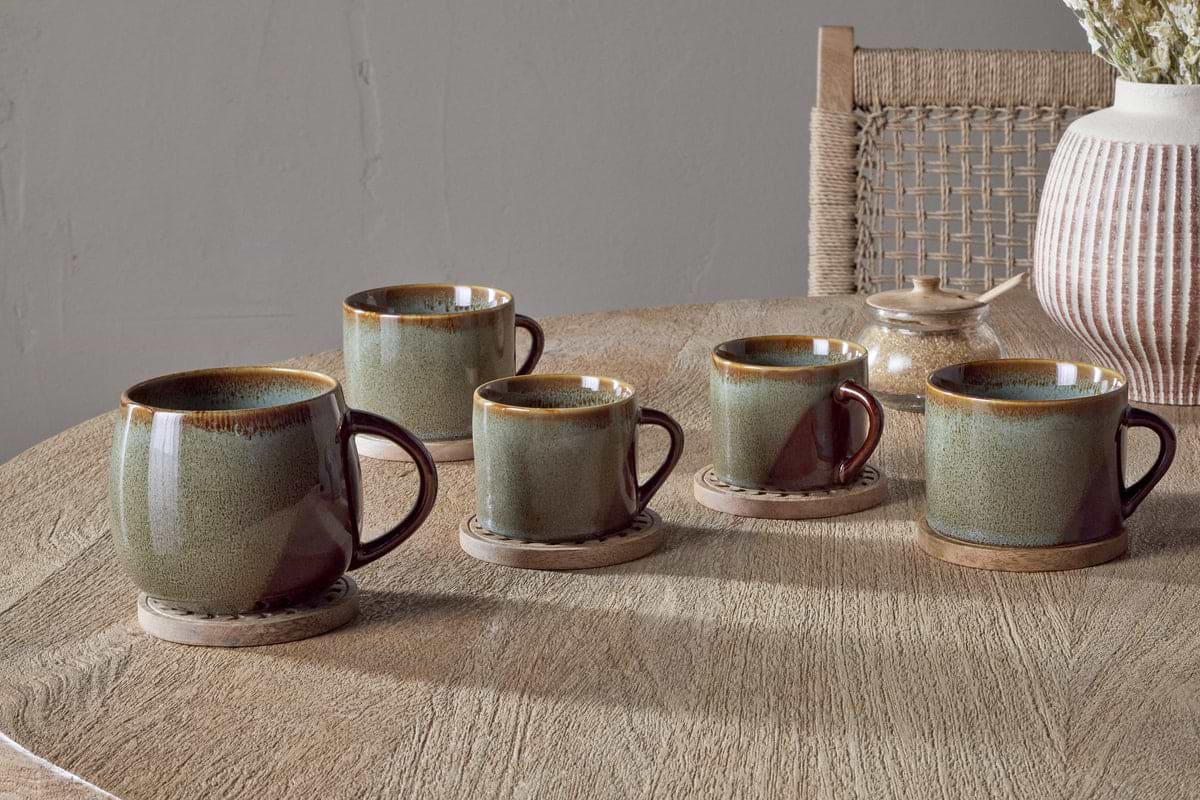 Two Naav Mugs - Blue - Large by nkuku, with a unique glaze effect, rest on wooden coasters atop a wooden table. In the background, a woven chair and dried flowers enhance the warm, rustic atmosphere.