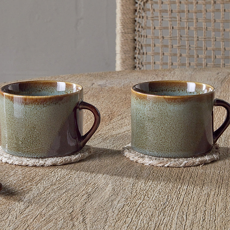 Two nkuku Naav Mugs - Blue - Large (Set of 2) with a unique glaze rest on woven coasters on a wooden table. A golden spoon with a brown handle and part of a textured vase enhance the distinctive tableware charm.