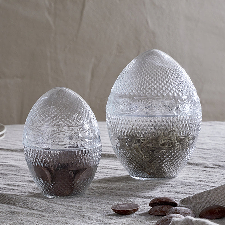 Two Essan Recycled Glass Opening Eggs by nkuku, made from clear textured recycled glass, sit on a light tablecloth. Each jar is filled with brown and green contents—perfect as unique decorative home accents. Plates are stacked left; a twig lies right.