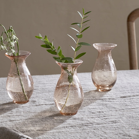 Three nkuku Bapti Bud Vases in Dusty Terracotta sit on a beige tablecloth. Two vases each hold a green sprig—one with drooping white flowers, one with leaves—while the third is empty. A wooden chair appears in the background.