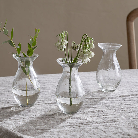 The nkuku Bapti Bud Vases - Clear (Set of 3) are displayed on a light gray linen tablecloth. One holds a leafy sprig, another has delicate white flowers, and the third is empty. A wooden chair can be seen in the background.