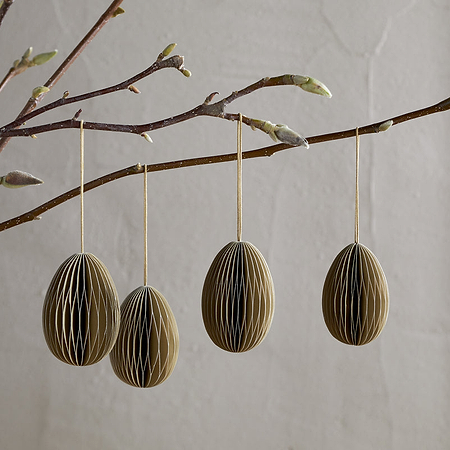 A budding branch in a gray vase displays four nkuku Piani Hanging Paper Decorations in moss green. The recycled ornaments hang on strings, set against a light, textured wall for a minimalist, natural spring look.