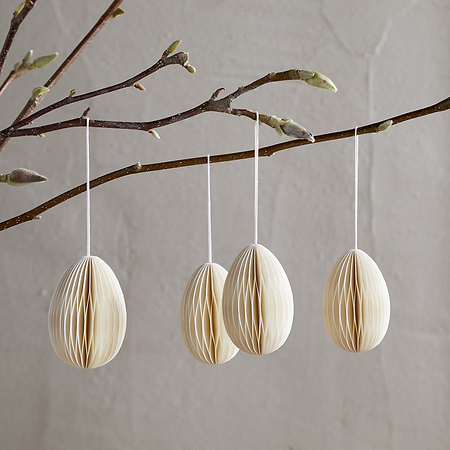 A ceramic vase holds a bare branch with buds, adorned with nkuku's Piani Hanging Paper Decoration - Cream (Set of 4), suspended by white strings. The recycled paper eggs stand out against a softly textured beige wall for a minimal, delicate look.