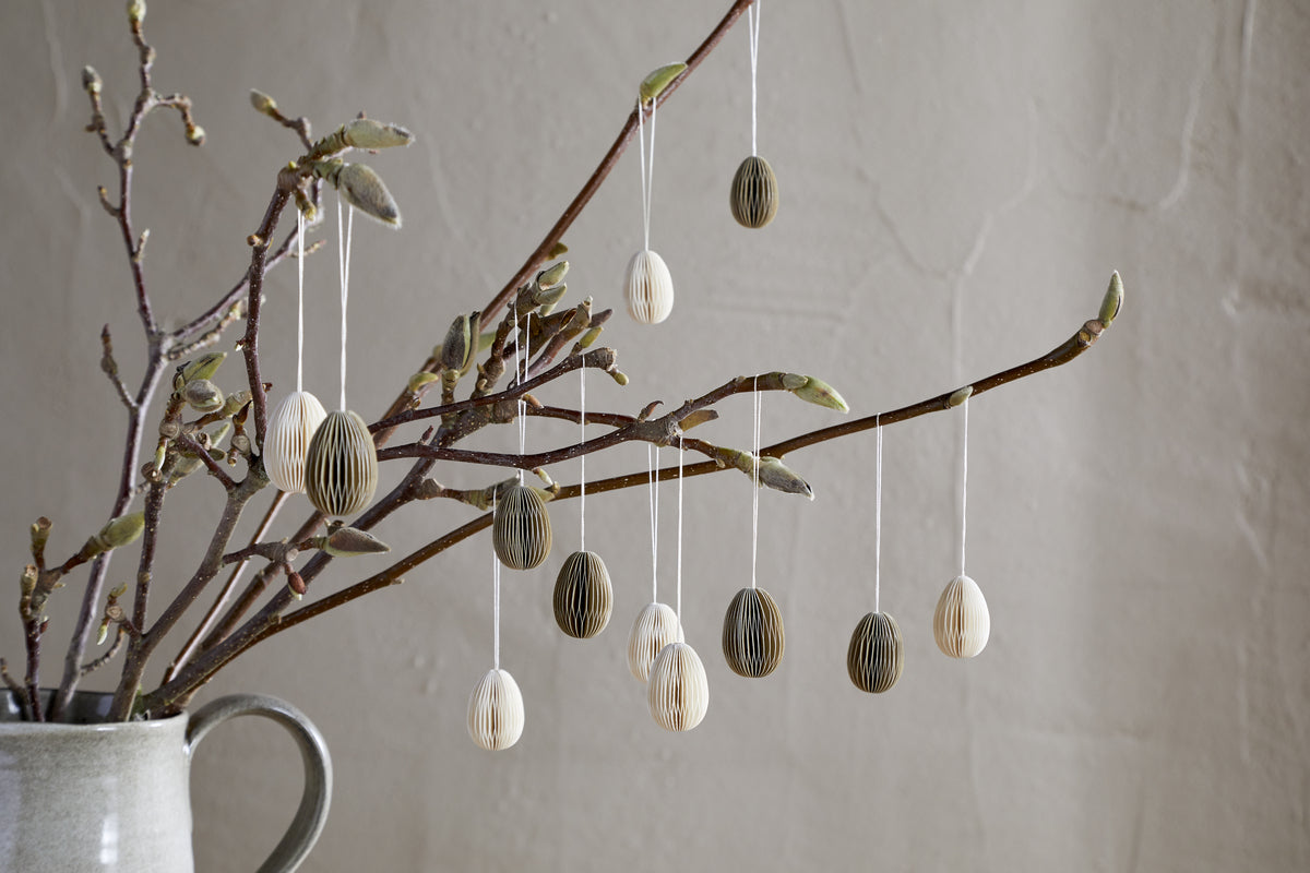 A ceramic jug holds budding branches, decorated with nkuku's Piani Mini Hanging Paper Decoration - Natural & Moss (set of 12), honeycomb eggs, and neutral accents, all set against a light wall for a calm, minimalist scene.