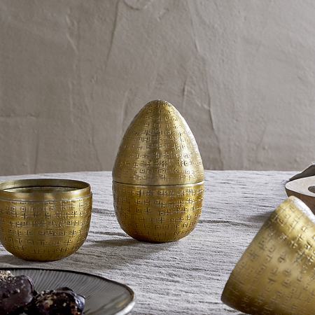 Three Mutti Hammered Opening Eggs in antique brass by nkuku, with intricate engraving, sit on a textured light gray tablecloth. One is open to reveal its interior. A plate of dark treats and a beige bowl with gray napkin are nearby.