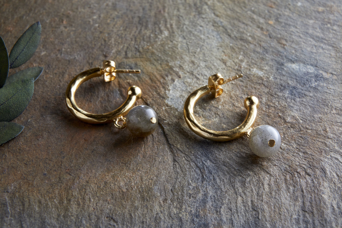 The Marai Labradorite Earrings by nkuku feature gold hoops with a hammered finish and elegant drop design, each adorned with a dangling round labradorite bead—a stylish jewellery gift.
