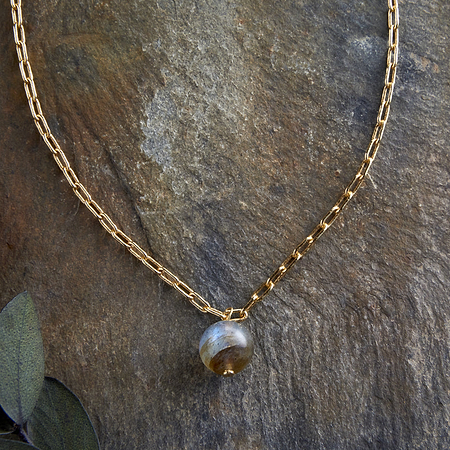 The Marai Labradorite Necklace - Labradorite & Gold by nkuku, featuring elongated gold links and a round marbled labradorite pendant, is displayed on brown stone with eucalyptus leaves—a perfect jewellery gift.