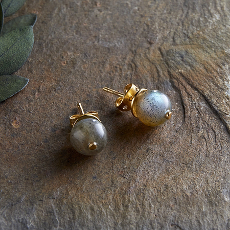 The Marai Labradorite Stud Earrings by nkuku feature round labradorite stones in gold settings, displayed on a textured stone surface with green leaves, highlighting the natural elegance and minimalist style of this jewelry.