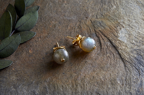 The Marai Labradorite Stud Earrings by nkuku feature round labradorite stones in gold settings, displayed on a textured stone surface with green leaves, highlighting the natural elegance and minimalist style of this jewelry.