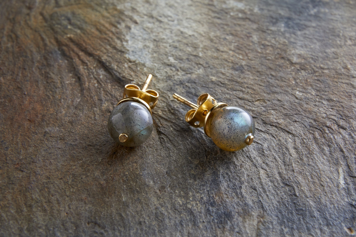 The Marai Labradorite Stud Earrings by nkuku feature round gray labradorite stones set in gold-tone metal, highlighting their natural variations—ideal for minimalist jewelry lovers.